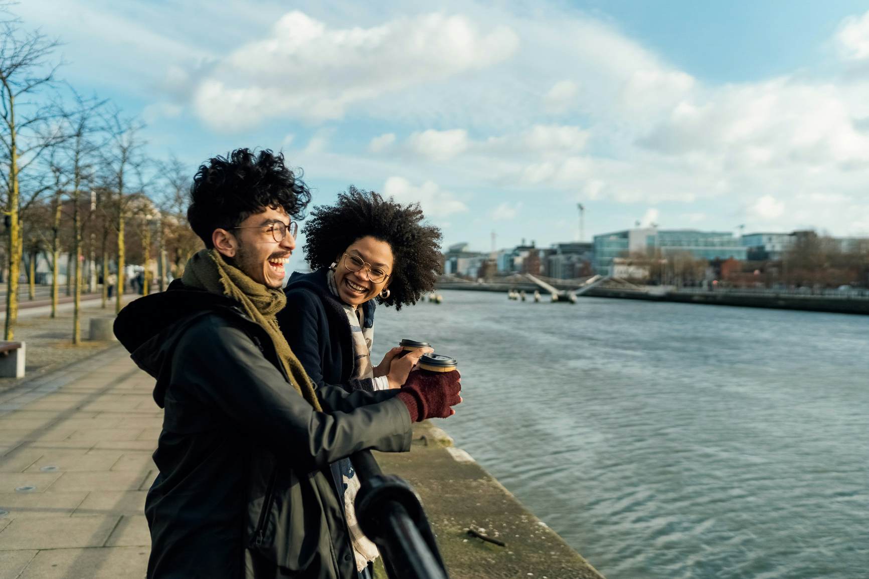 Young Couple of Friends Are meeting in Town for a takeaway Coffee a Walk by the river. The urban area is lighted by sunny cold autumn/ winter day. the Latin Man and the Afro Black Woman are Wearing Heavy jackets and holding a Cup of Coffee from the Shon in The Corner.